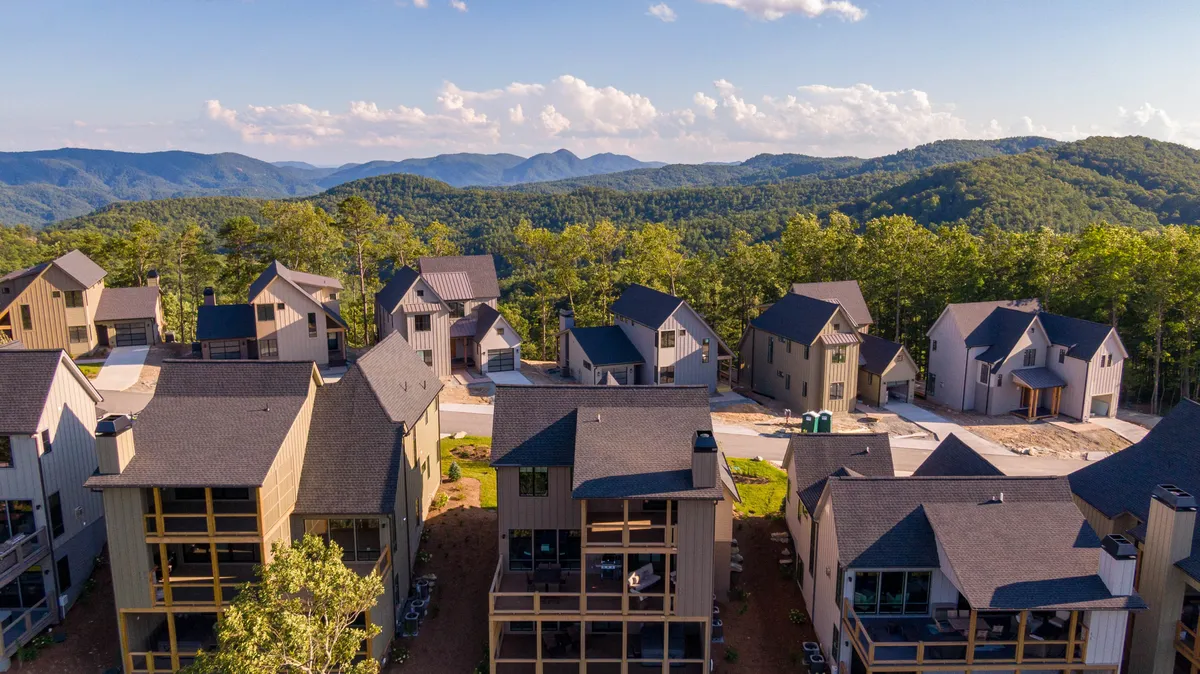 The image showcases a modern mountain home nestled in lush greenery, with expansive views of the Blue Ridge Mountains stretching into the distance. The home's architecture is sleek and inviting, and the elevated setting hints at the serenity and privacy guests can expect. Evening light bathes the property, suggesting peaceful outdoor relaxation and opportunities for stargazing.