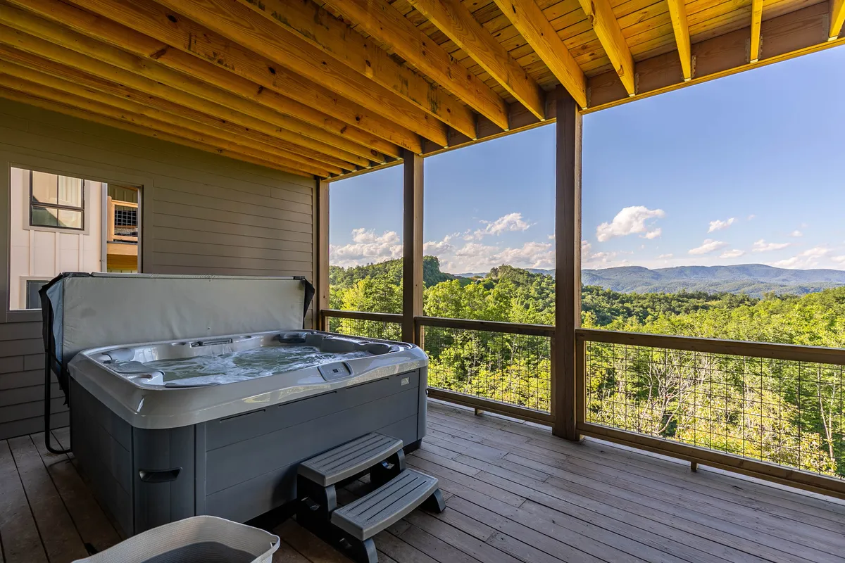 A steaming hot tub sits on a covered deck with a panoramic view of rolling mountains. The tranquil setting invites relaxation after a day of outdoor adventures, perfectly blending luxury and nature.