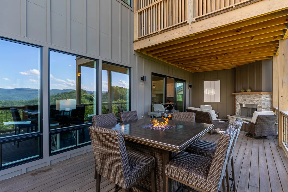 An expansive exterior deck showcases a modern dining table with a built-in fire feature and plush seating. The space opens to breathtaking mountain vistas, with large glass windows connecting indoor and outdoor living.